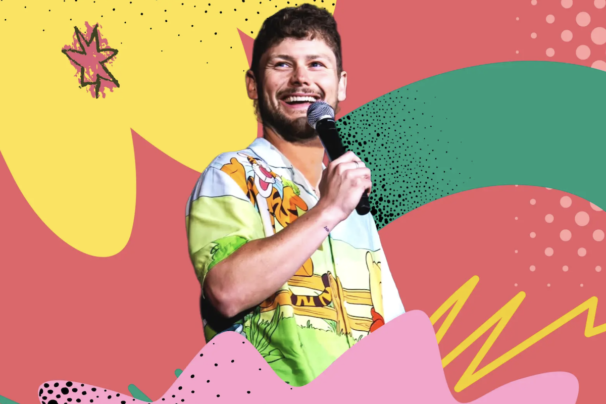 Comedian Drew Lynch smiling on stage into a microphone in front of a colorful pop-art background.